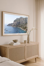 Light oak framed Capri photography print styled above a light wood cabinet in a coastal bedroom interior.
