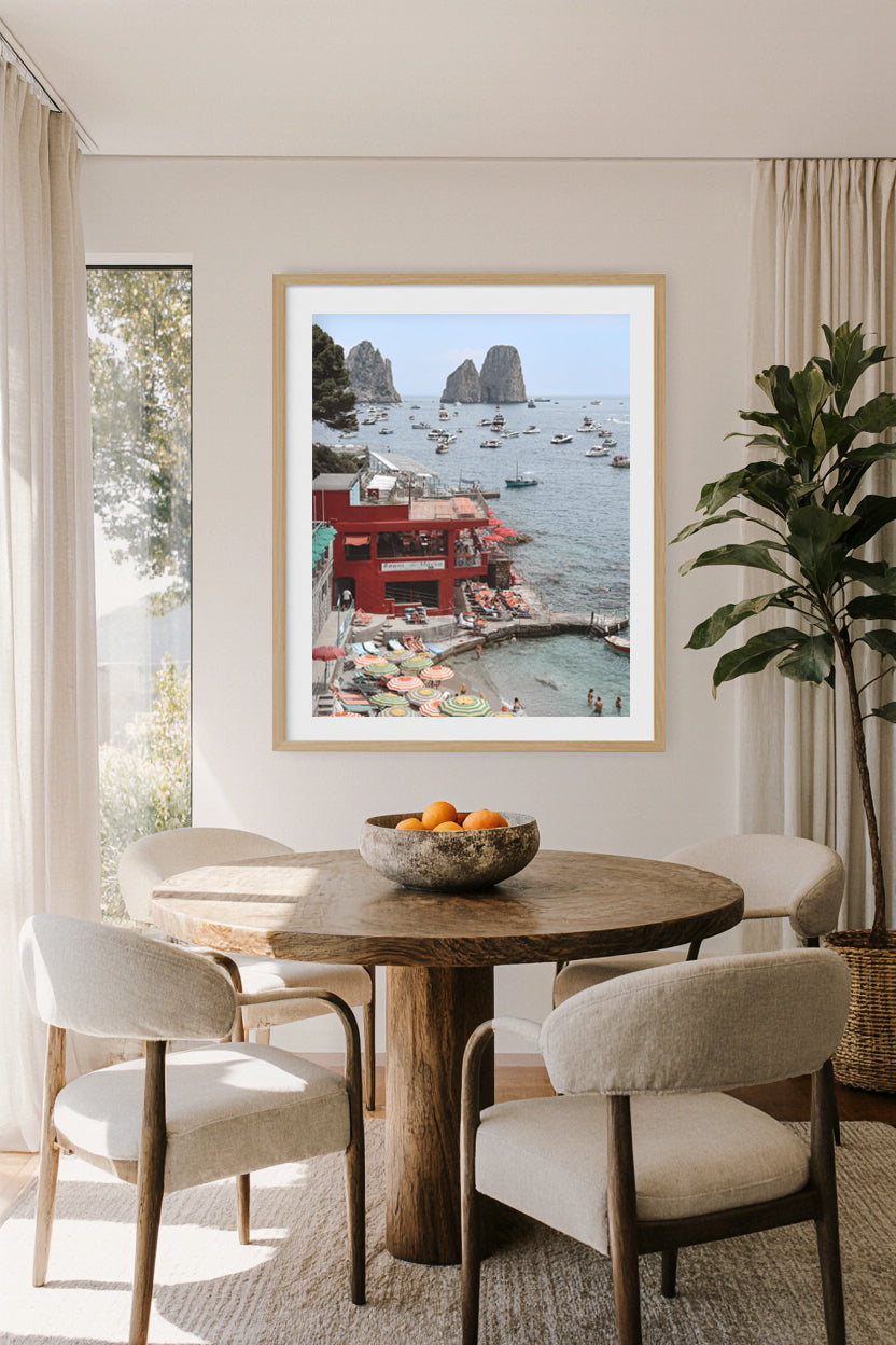 Dining room with a round wooden table and chairs, featuring a framed picture of Marina Piccola beach and the Faraglioni Rock in Capri, Italy on the wall.