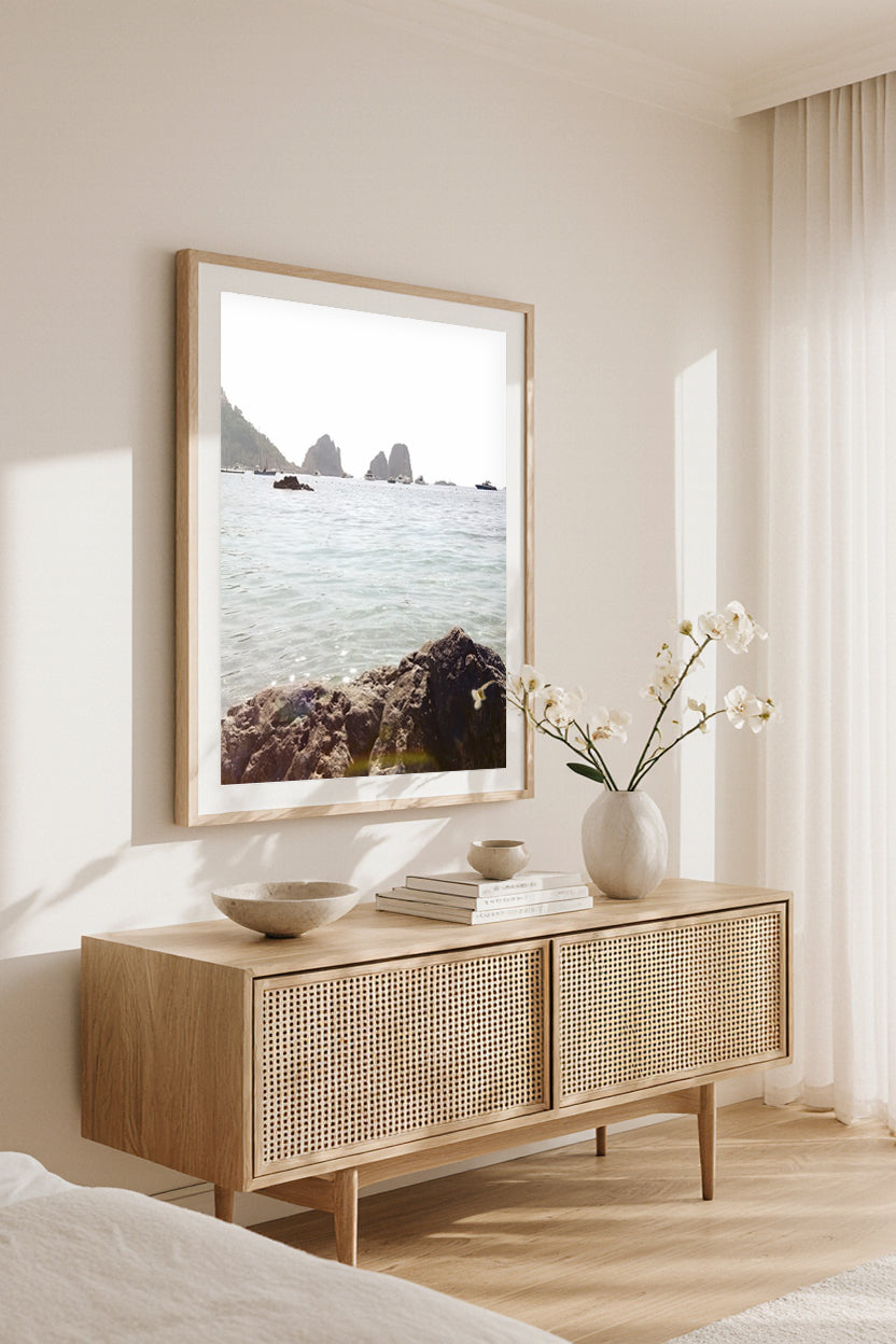 Framed wall art print of Capri’s Faraglioni sea stacks, styled in a coastal bedroom above a natural wood console with orchids.