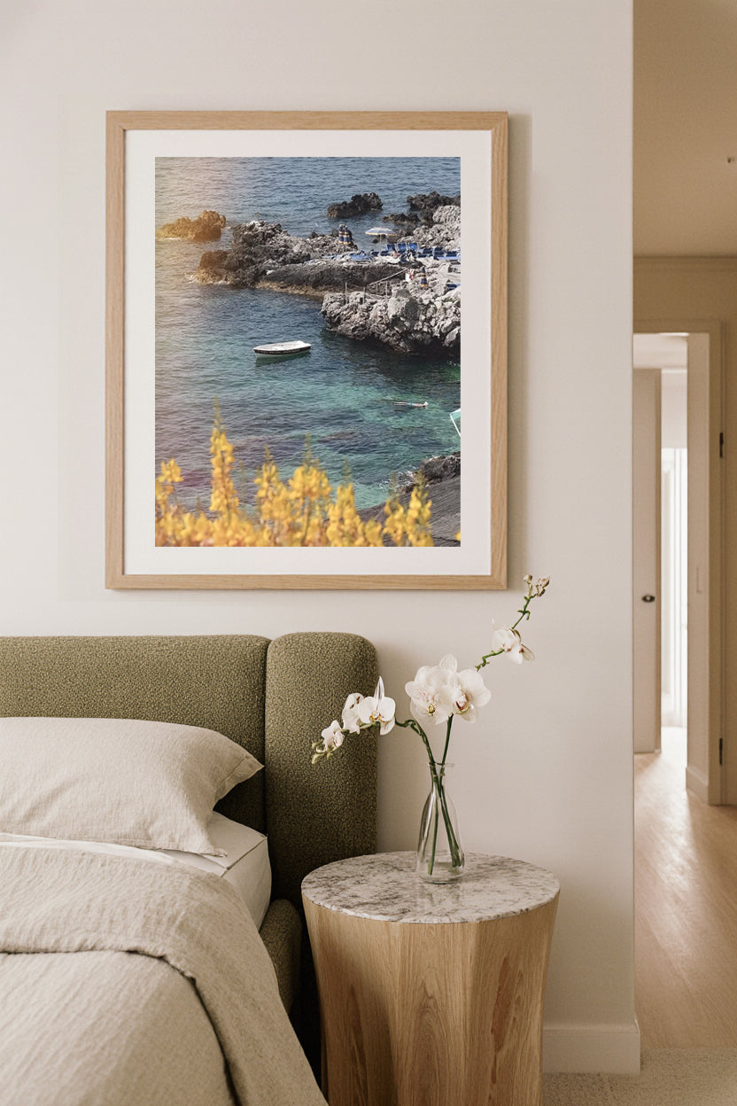 Framed artwork of a coastal scene above a bed in a modern bedroom. Coastal travel photography bringing Italian summers to home décor.