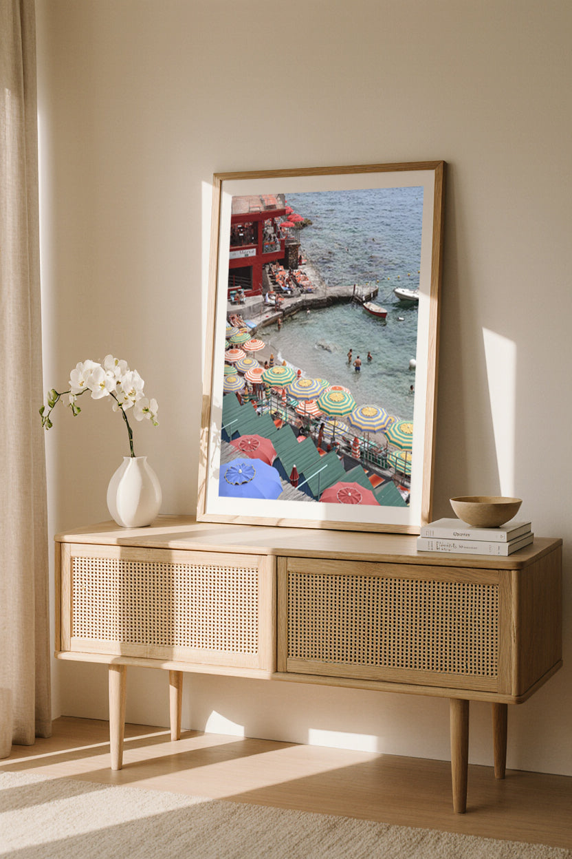Framed artwork of Marina Piccola Beach Club on a wooden console table. Capri travel photography with striped umbrellas and turquoise sea, elegant coastal wall art to elevate modern luxury home décor.