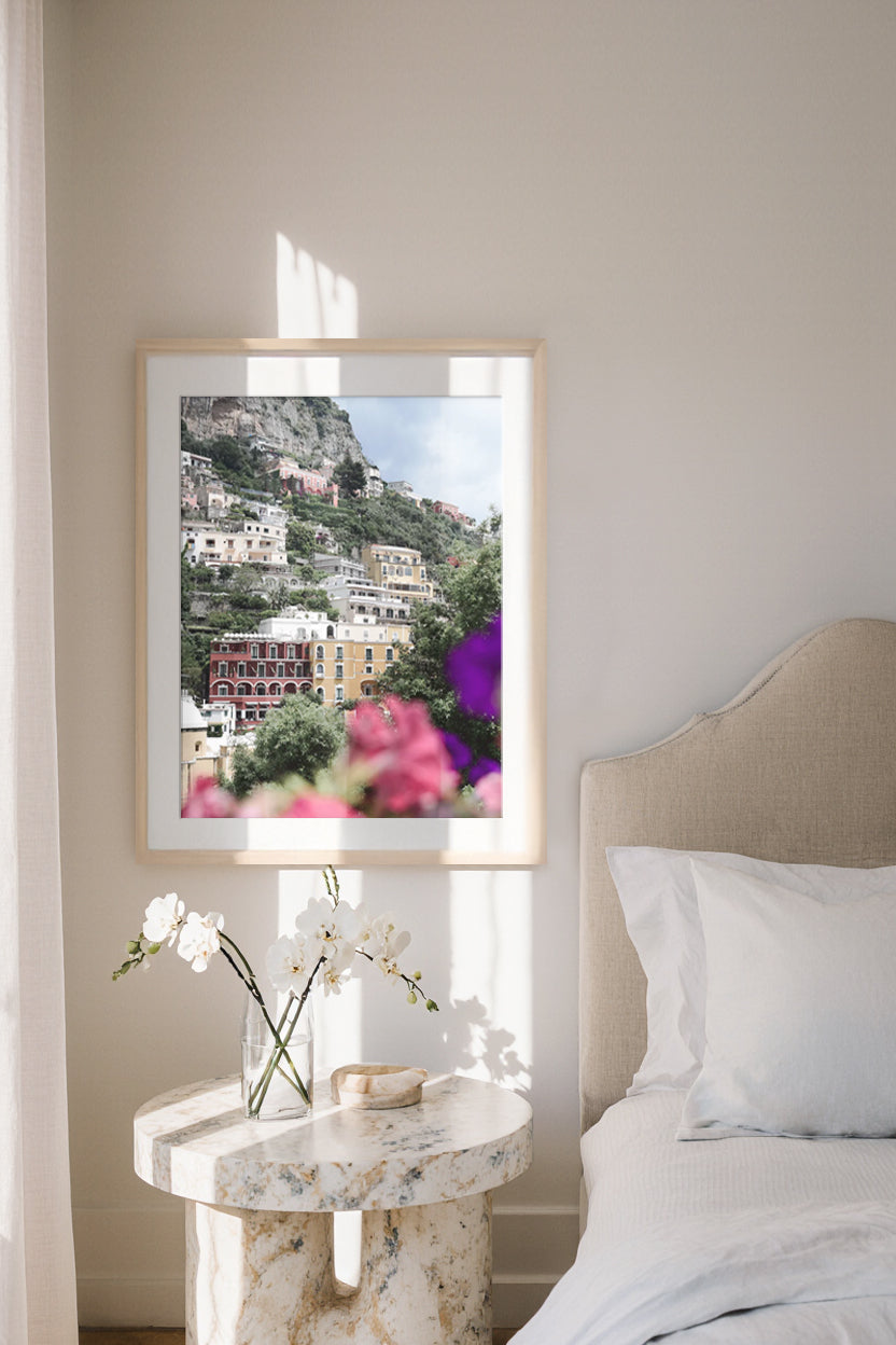Framed Positano artwork of a scenic pastel landscape on a wall above a marble nightstand with flowers.