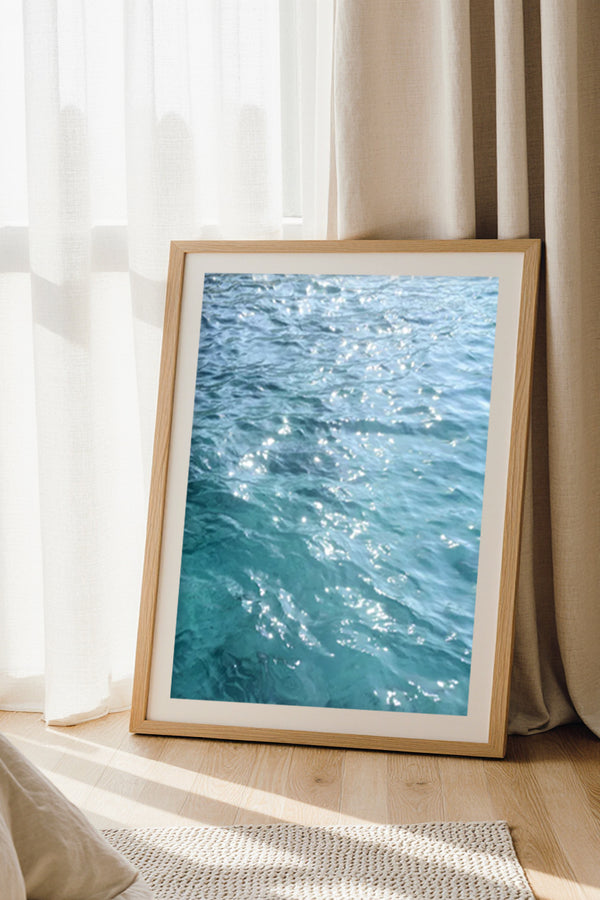 Fine art photography print of sparkling turquoise Mediterranean water, framed wall art styled on the floor by a window with curtains.