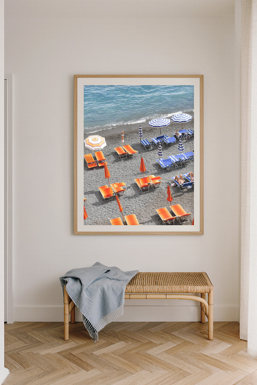 Fine art photography print of Positano beach club in Italy, featuring vibrant orange sunbeds and blue striped umbrellas along the shoreline, coastal Mediterranean wall art décor.