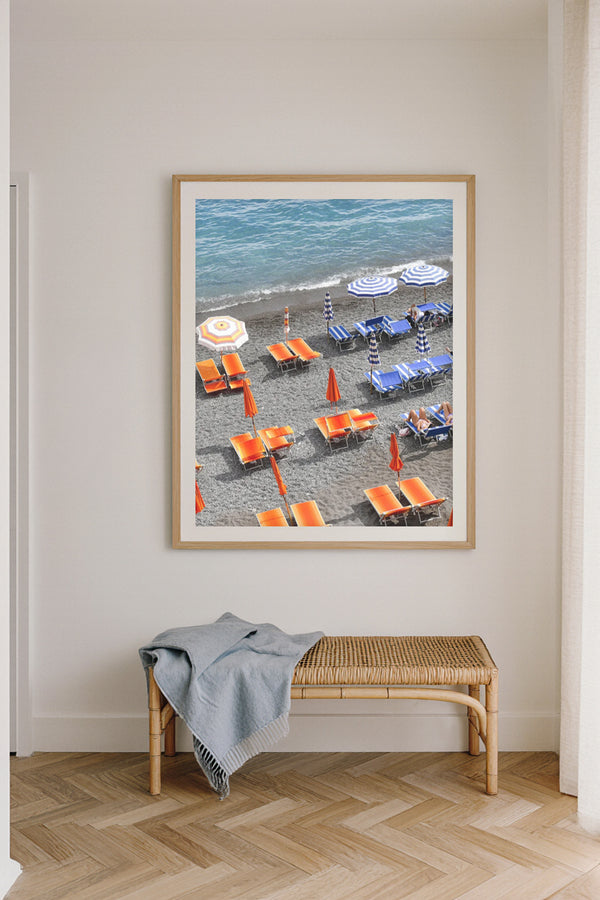 Fine art photography print of Positano beach club in Italy, featuring vibrant orange sunbeds and blue striped umbrellas along the shoreline, coastal Mediterranean wall art décor.