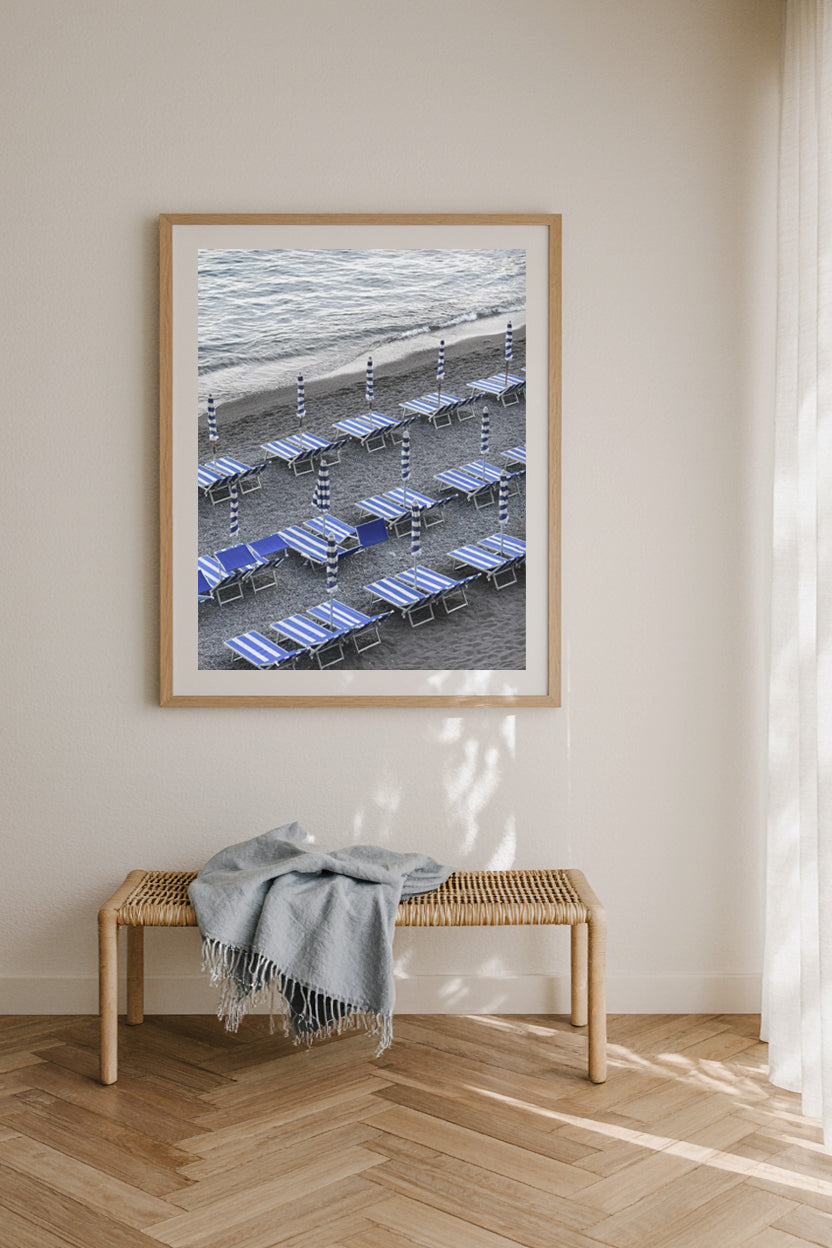 Framed artwork of a beach scene with lounge chairs on a wall above a wooden bench.