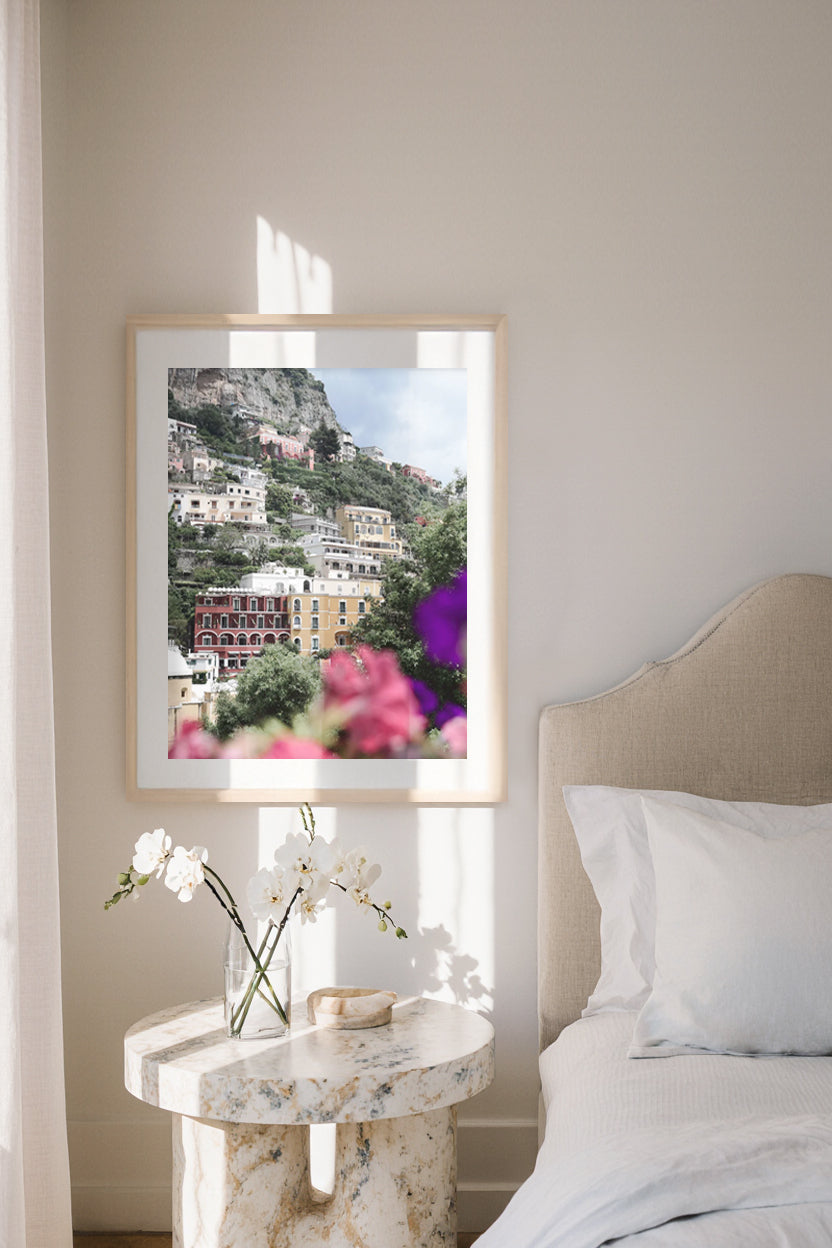 Framed Positano artwork of a scenic pastel landscape on a wall above a marble nightstand with flowers.